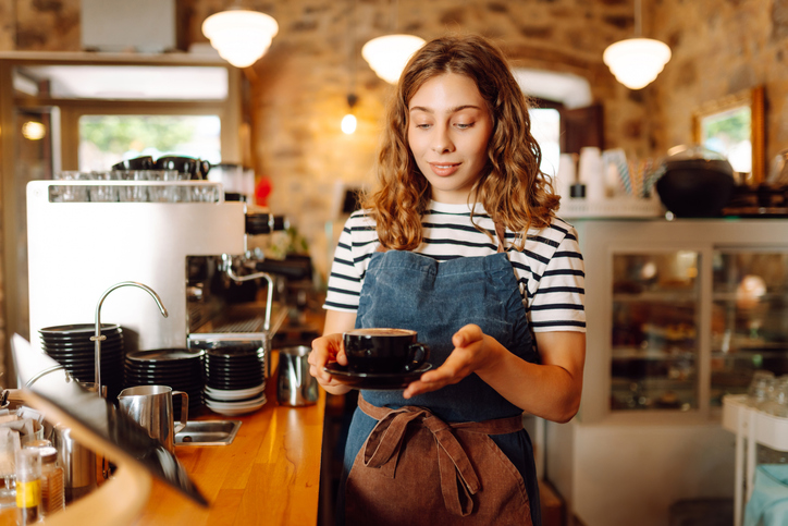 Happy female barista holding a cup of hot and aromatic coffee in her hands standing behind the bar. Young employee of the coffee shop gives coffee to a customer. Concept of drinks, small business.